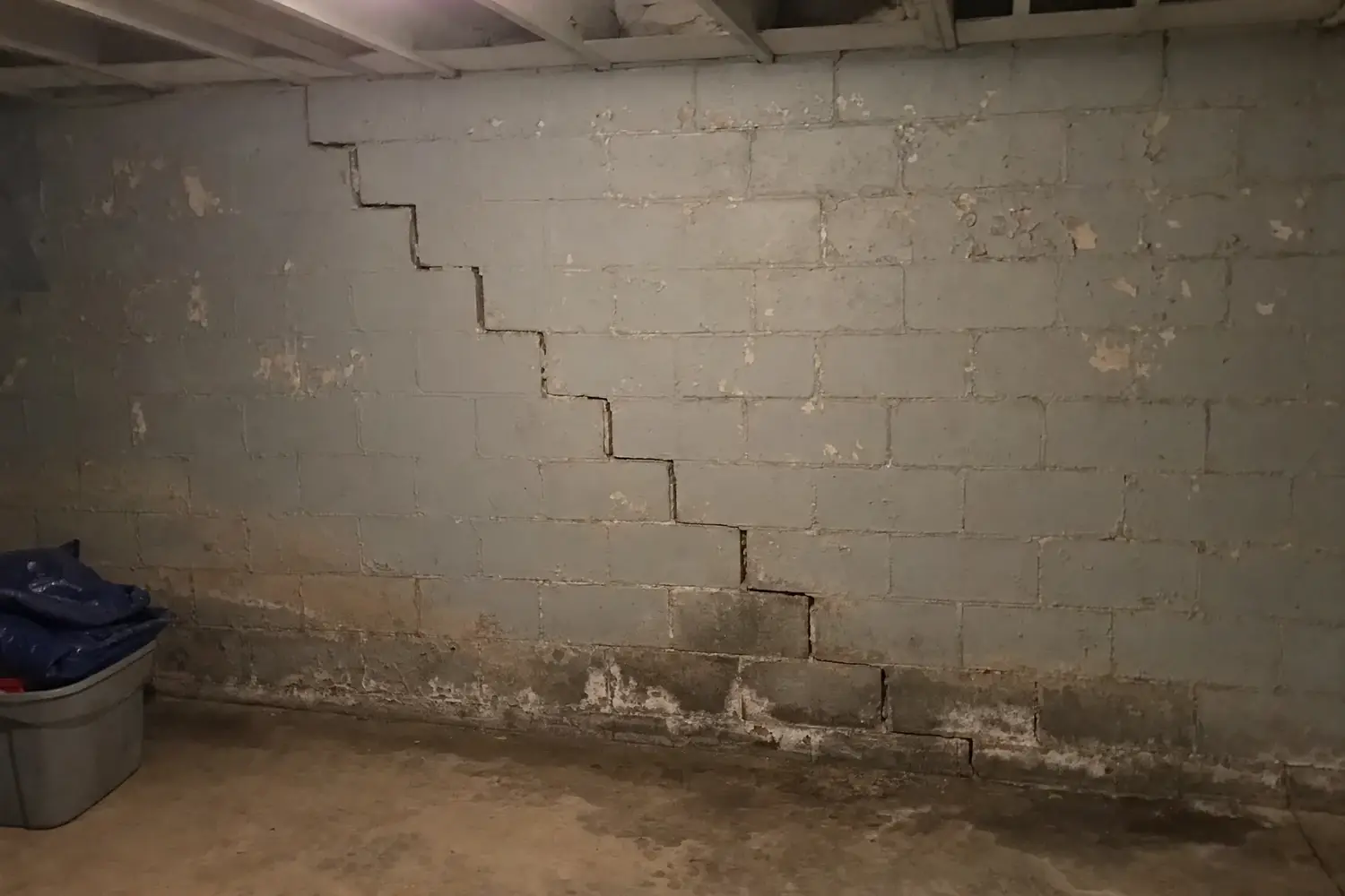 Stair-step crack running down the mortar joints of a painted concrete block basement wall, with water-stained lower courses marked by white efflorescence