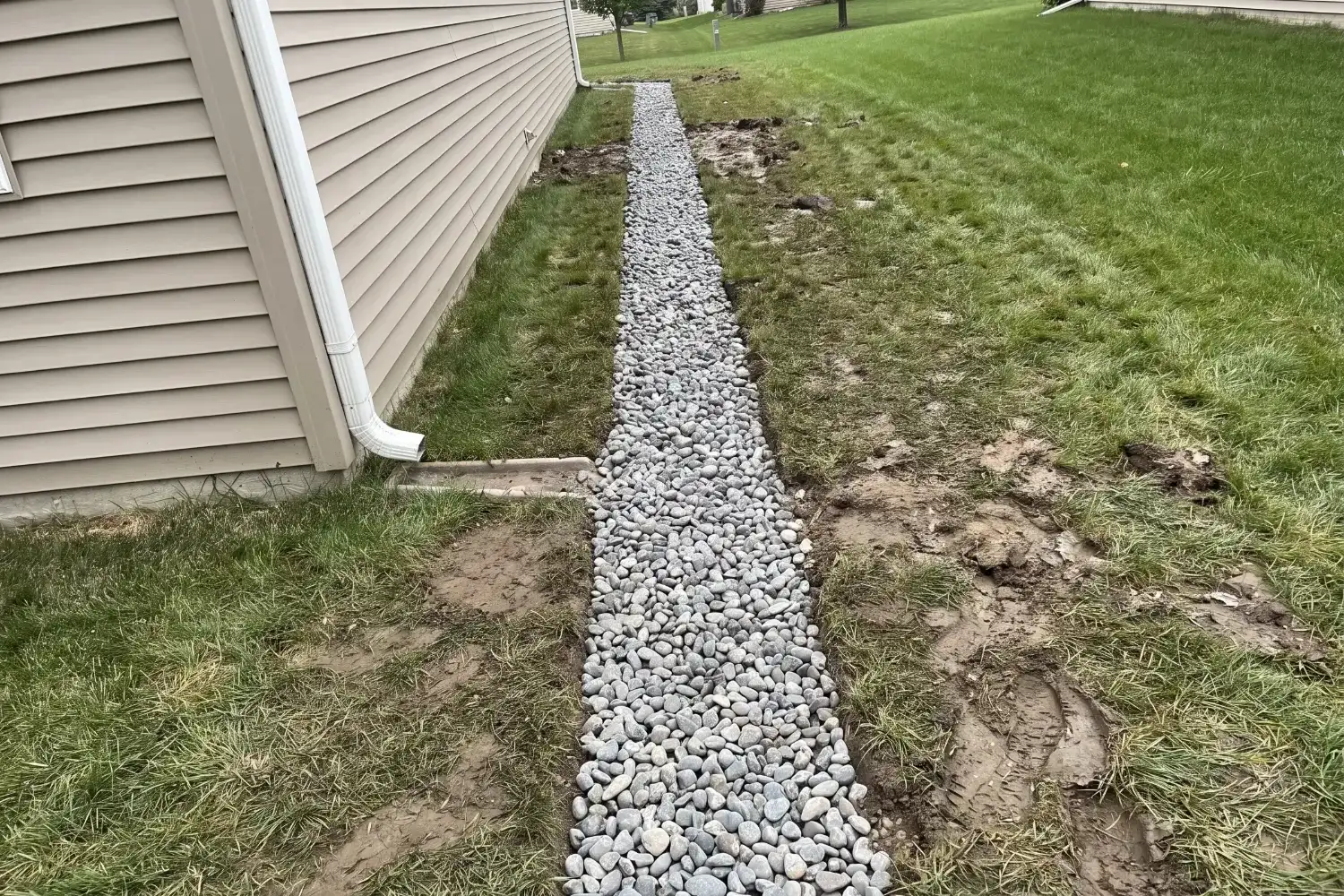 Newly installed French drain trench filled with river rock, running through a side yard away from a downspout