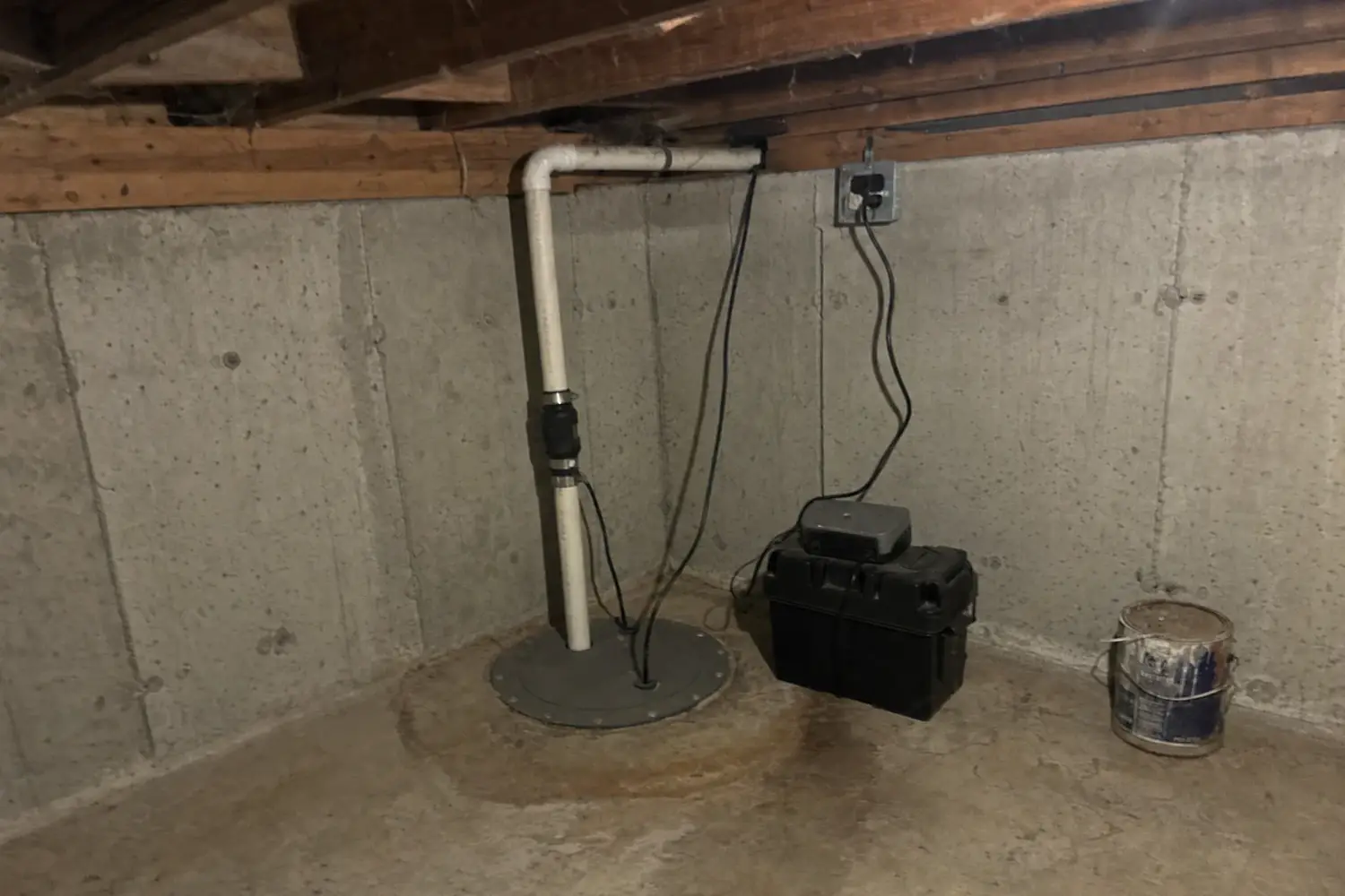 Sealed, bolted sump pit lid with a white PVC discharge pipe and rubber check valve, next to a battery backup box with its charger on top, in a poured concrete corner under the floor joists