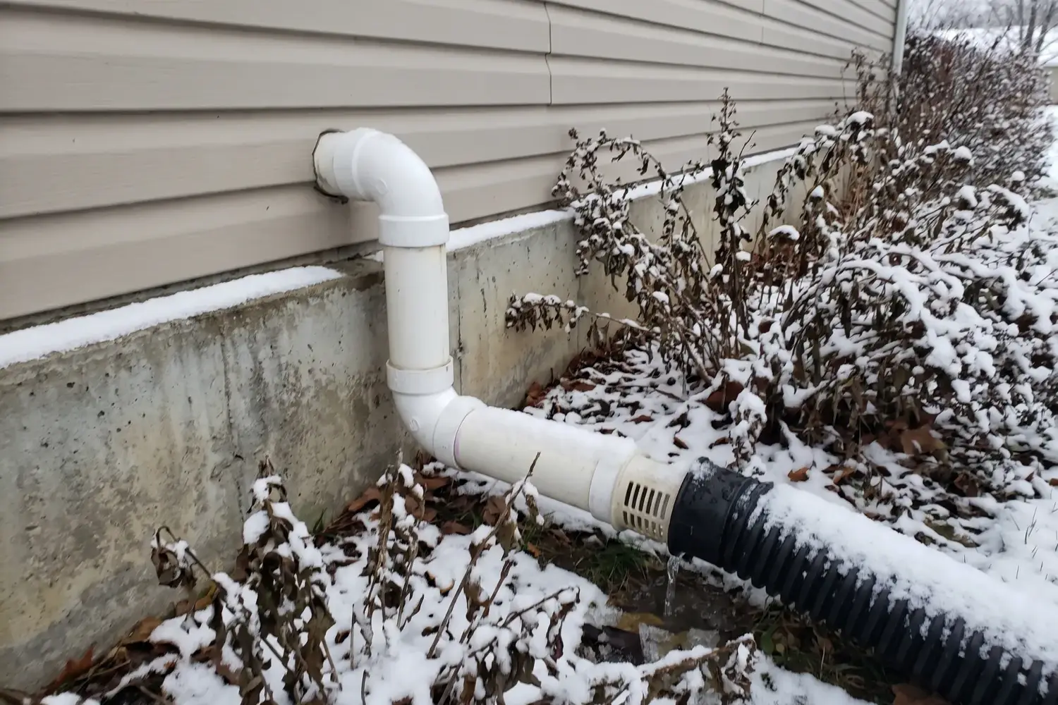Sump pump discharge line exiting a house above the foundation and draining through a corrugated extension pipe in winter snow